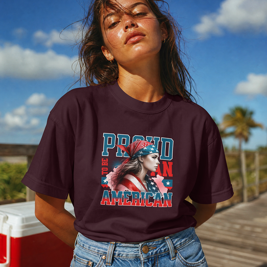 A woman outdoors wearing a dark maroon graphic T-shirt with the text 'PROUD TO BE AMERICAN' and a patriotic illustration, paired with blue jeans. The background shows a sunny beach or boardwalk scene with blue sky, clouds, and palm trees.